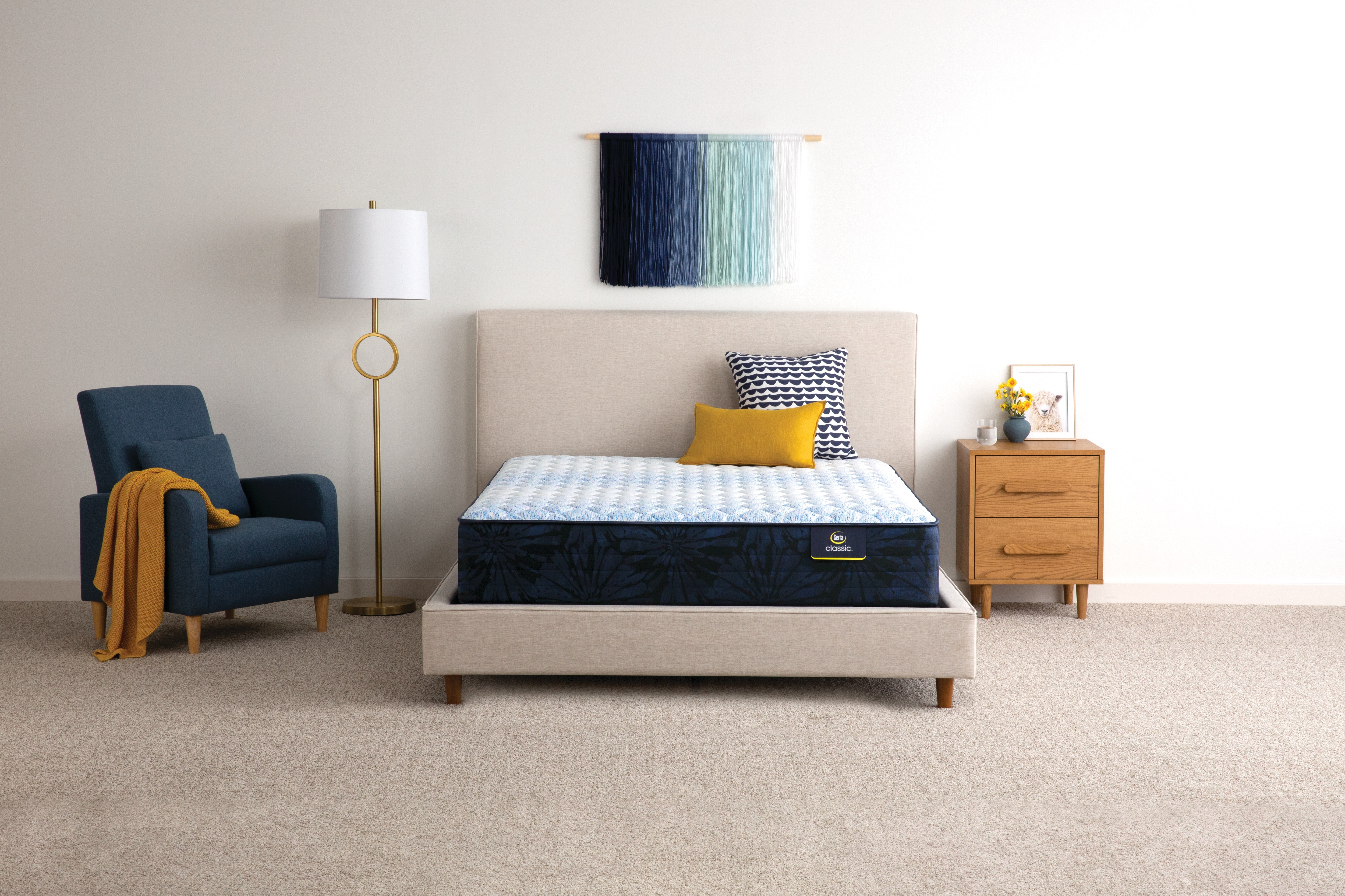 Serta Octave firm tight top mattress in modern room sitting on beige platform with headboard, wooden side table and blue chair with brown throw blanket